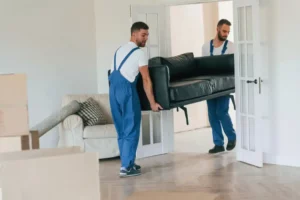 Zwei Umzugshelfer in blauen Latzhosen tragen ein schwarzes Ledersofa durch eine Flügeltür