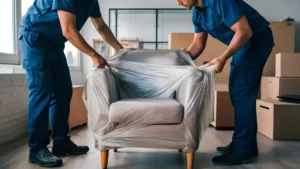 Zwei Umzugshelfer in blauer Arbeitskleidung wickeln einen Sessel in Stretchfolie ein