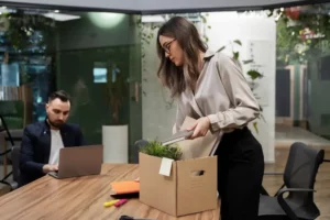 Frau packt im Büro Ordner und eine Pflanze in einen Umzugskarton, ein Kollege arbeitet daneben am Laptop